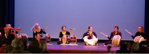 Group of performers seated on stage in a drum circle, playing large frame drums while the audience watches from the front row.