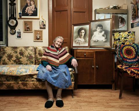swifthouseinn_hotel_middlebury_hirschfieldfilmseries Older woman sits on a floral sofa in a cozy living room, surrounded by framed family photos on the wall and cabinet.