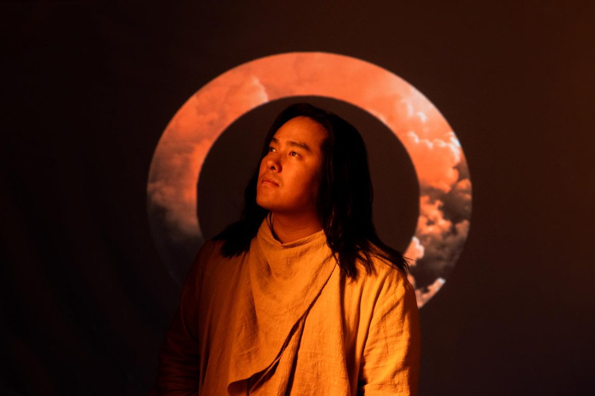 Person with long dark hair in orange robes gazes upward, standing before a large orange circular projection resembling a halo of clouds.