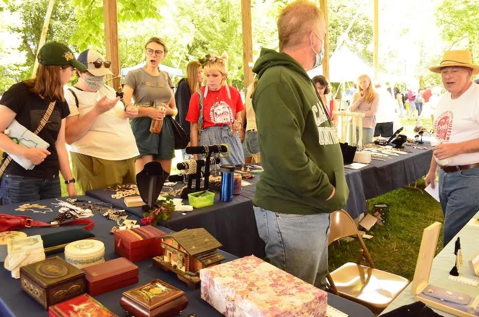 Group of people browsing a sunny outdoor craft market; tables overflow with boxes, jewelry, and small crafts.