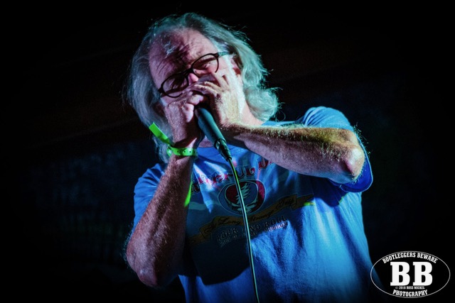 Older man with gray hair and glasses singing into a handheld microphone on a dark stage, wearing a blue T‑shirt.