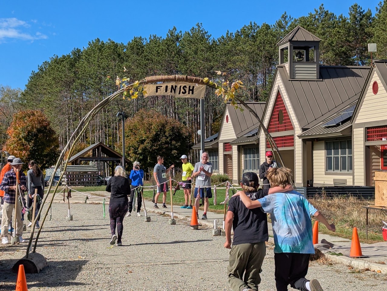 Runners approach the finish line under a curved arch, a banner reading FINISH, while spectators cheer on a sunny gravel path near a beige building.