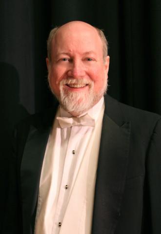 Smiling older man with a white beard in a black tuxedo and white bow tie, standing against a dark curtain backdrop.